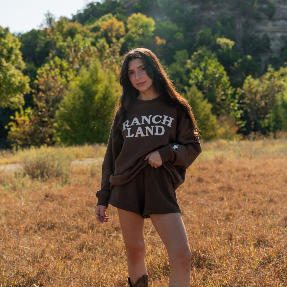 Full-length photo of a model in a golden field wearing the brown Ranch Land Essentials sweatshirt and matching shorts with leather cowboy boots.