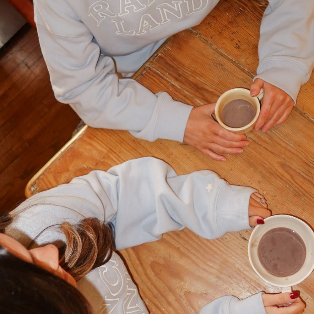Overhead view of two people in light blue Misty collection sweatshirts sitting at a rustic wooden table with mugs of coffee.
