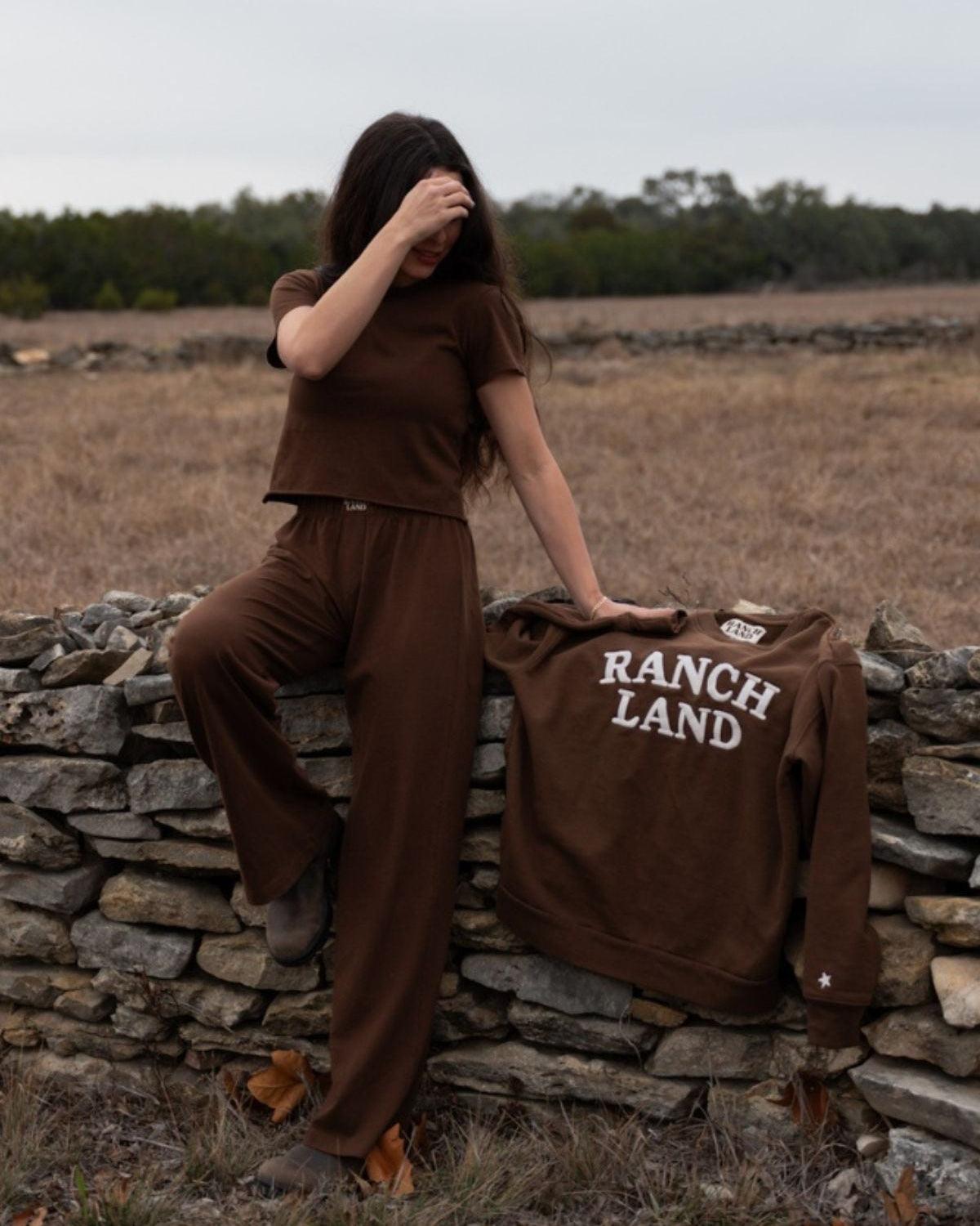 Person in brown outfit sitting on a stone wall with 'Ranch Land' shirt in a field.