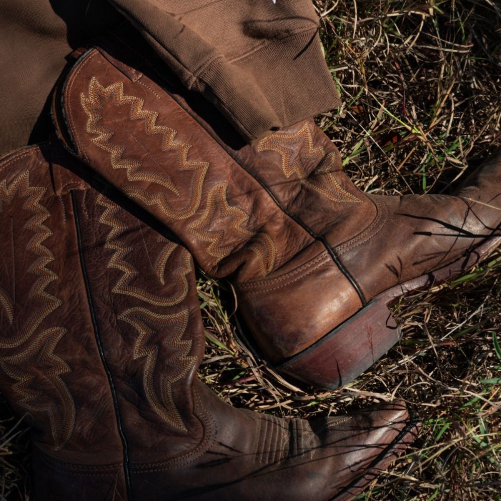 Close-up of worn brown leather cowboy boots with intricate stitching lying in dry grass next to a brown organic cotton sweatshirt sleeve.