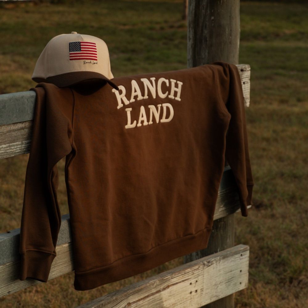 A brown Ranch Land sweatshirt with cream 'RANCH LAND' back text draped over a wooden ranch fence next to a tan baseball cap featuring an American flag.