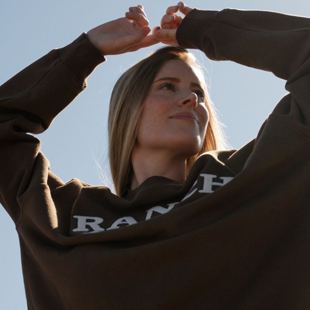 Low-angle sunlit portrait of a blonde woman wearing a brown Ranch Land organic cotton sweatshirt against a clear blue sky.
