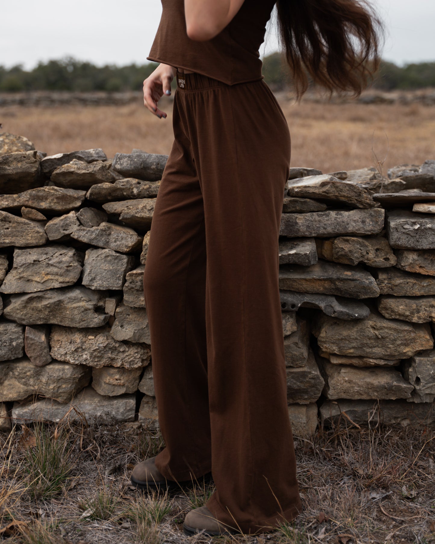 Person wearing brown pants standing in a field with a stone wall in the background