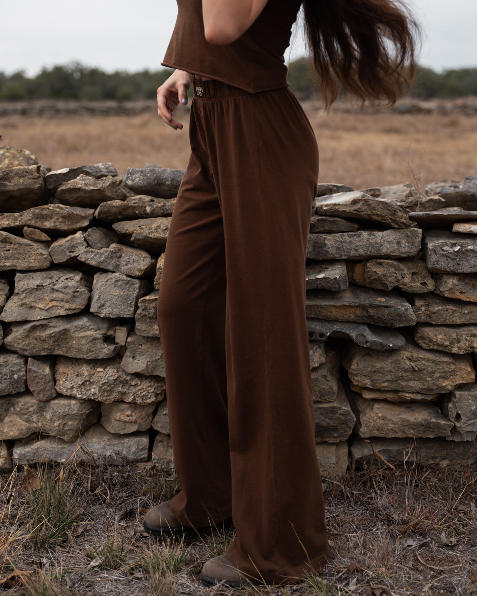 Person wearing brown pants standing in a field with a stone wall in the background
