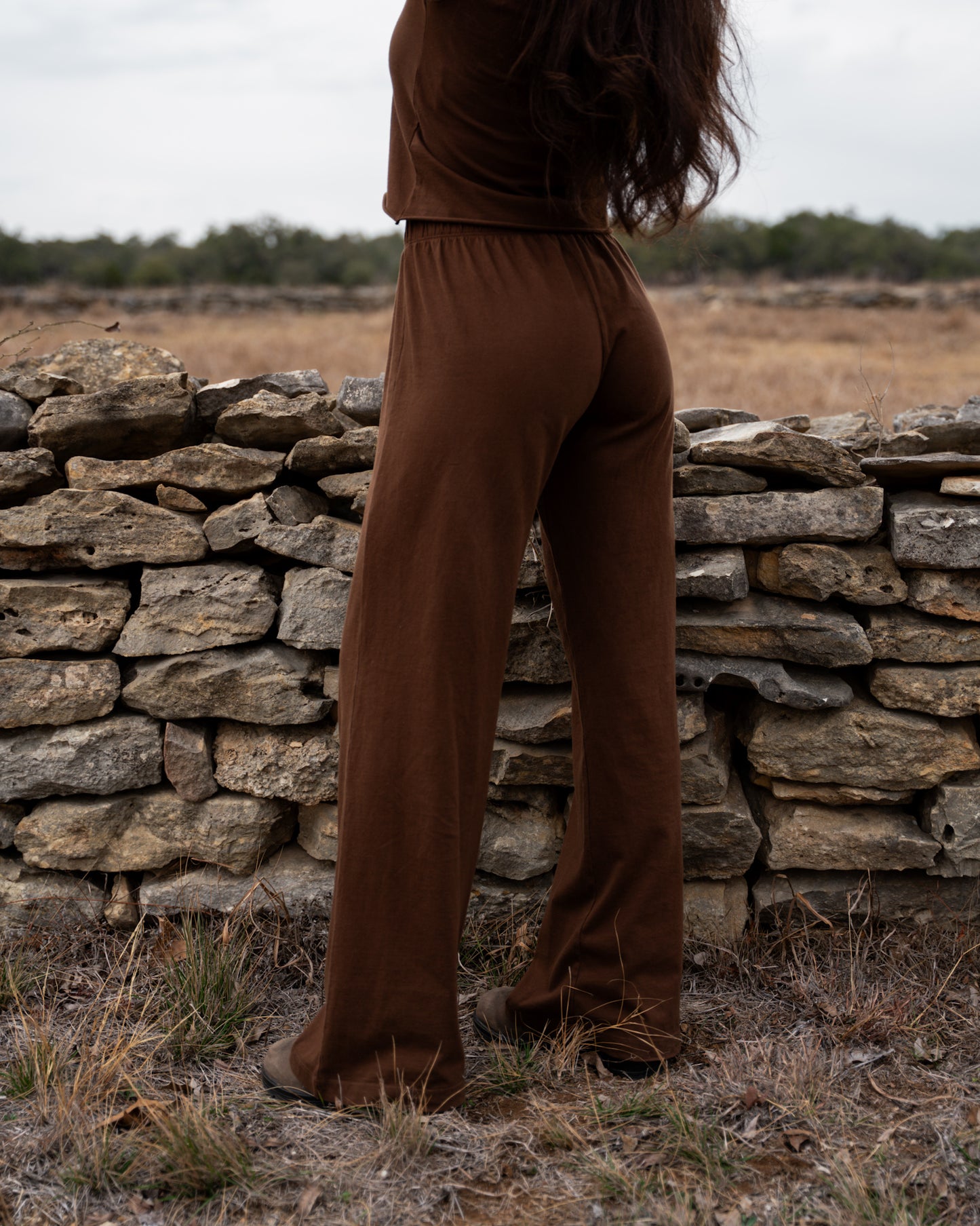 Person wearing brown pants standing in a field with a stone wall in the background