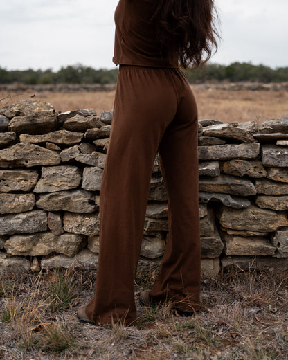 Person wearing brown pants standing in a field with a stone wall in the background