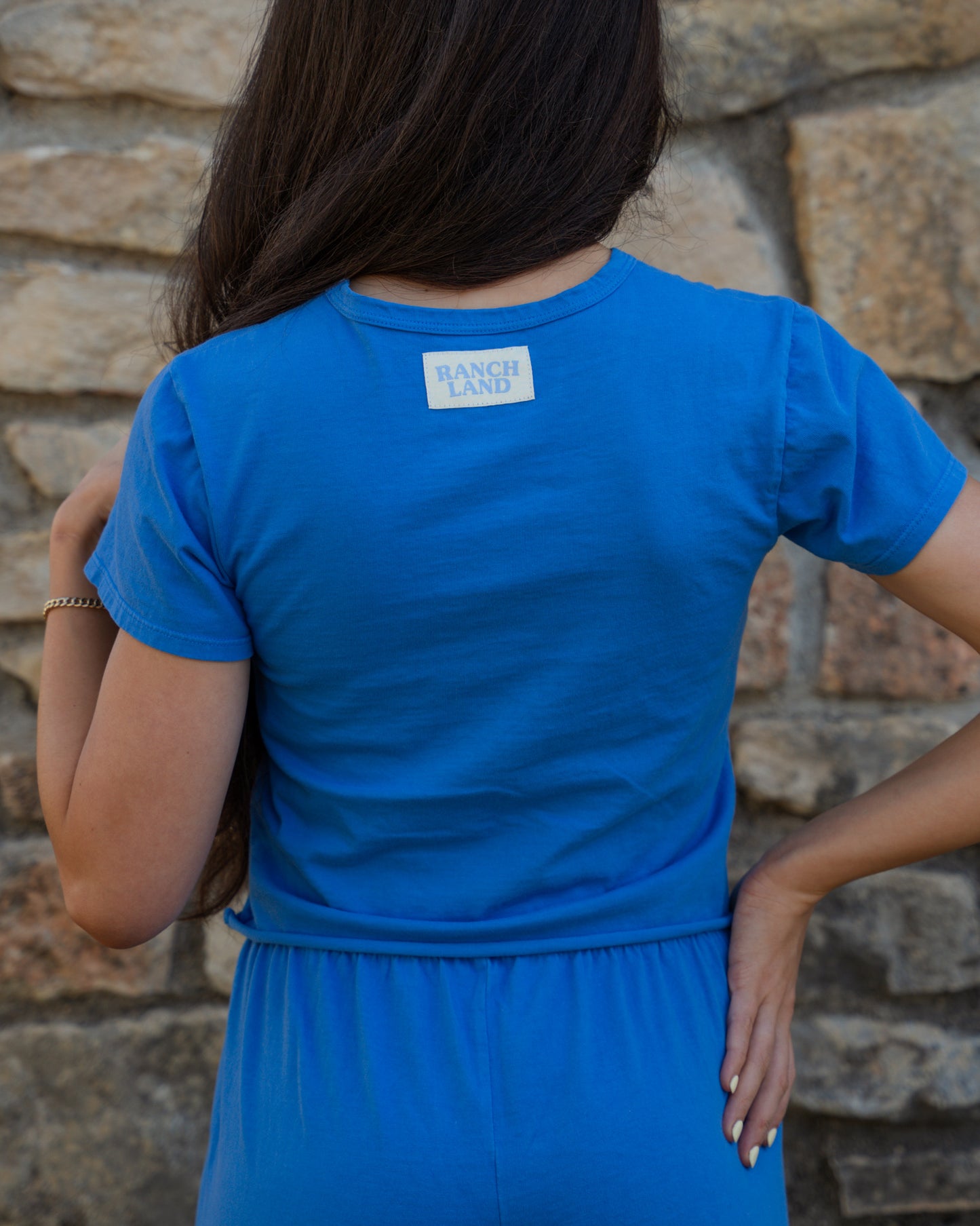 Person wearing a blue t-shirt with text on the back against a stone wall.
