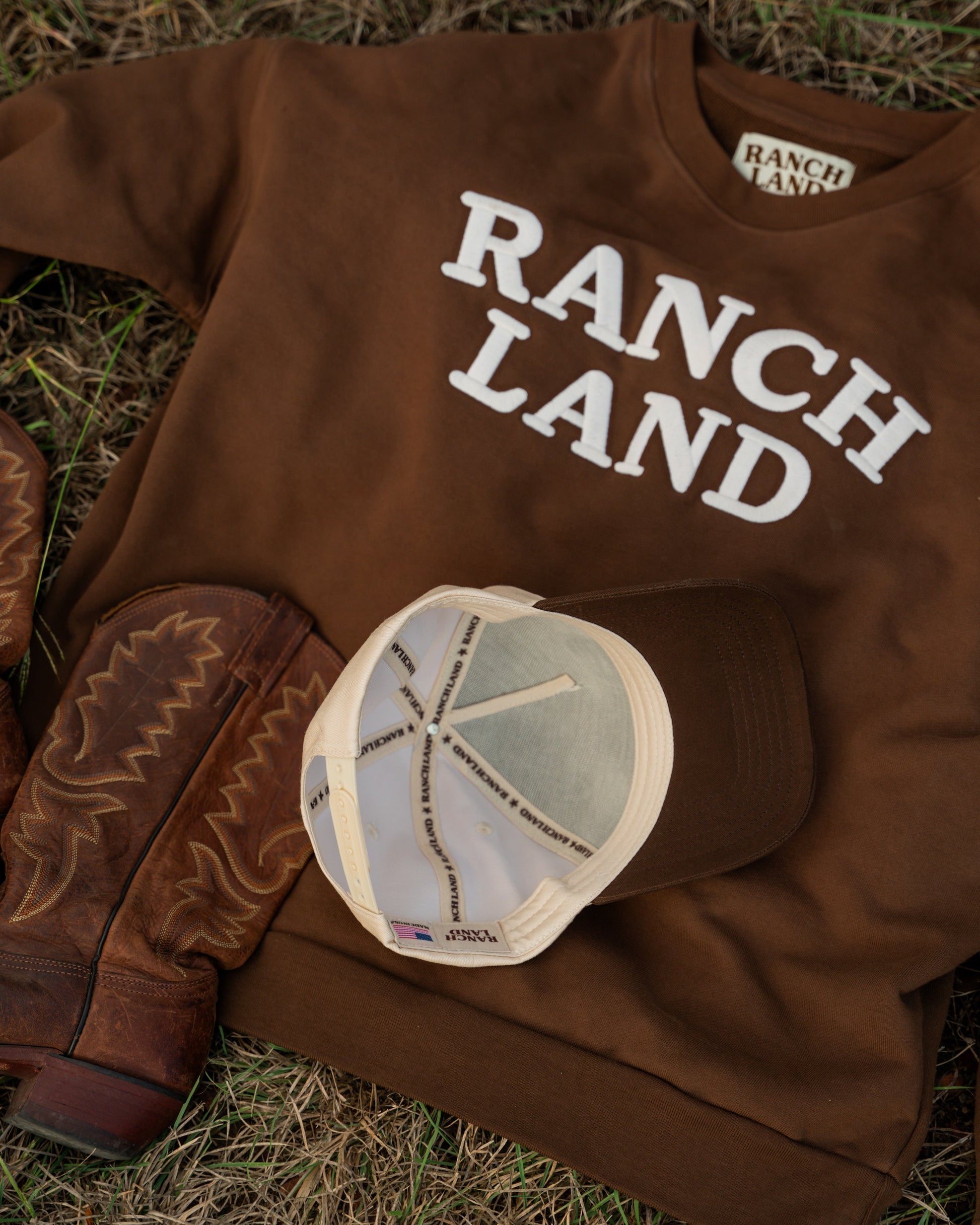 Brown 'Ranch Land' sweatshirt with cowboy boots and a cap on grass