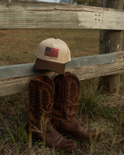 Brown cowboy boots and cap with American flag design leaning against a wooden fence in a field.