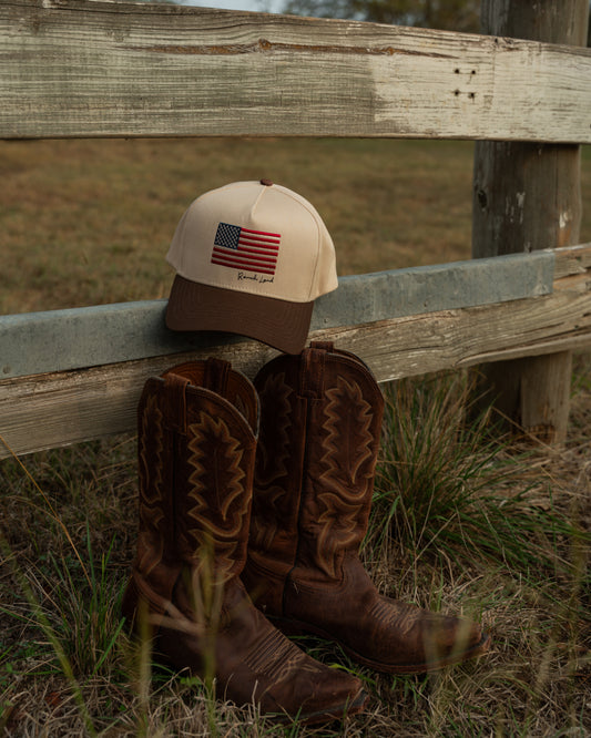 Brown cowboy boots and cap with American flag design leaning against a wooden fence in a field.