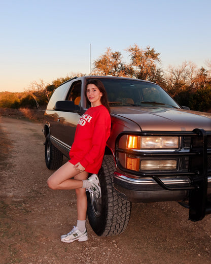 Person in a red sweatshirt leaning against a brown SUV on a dirt road.