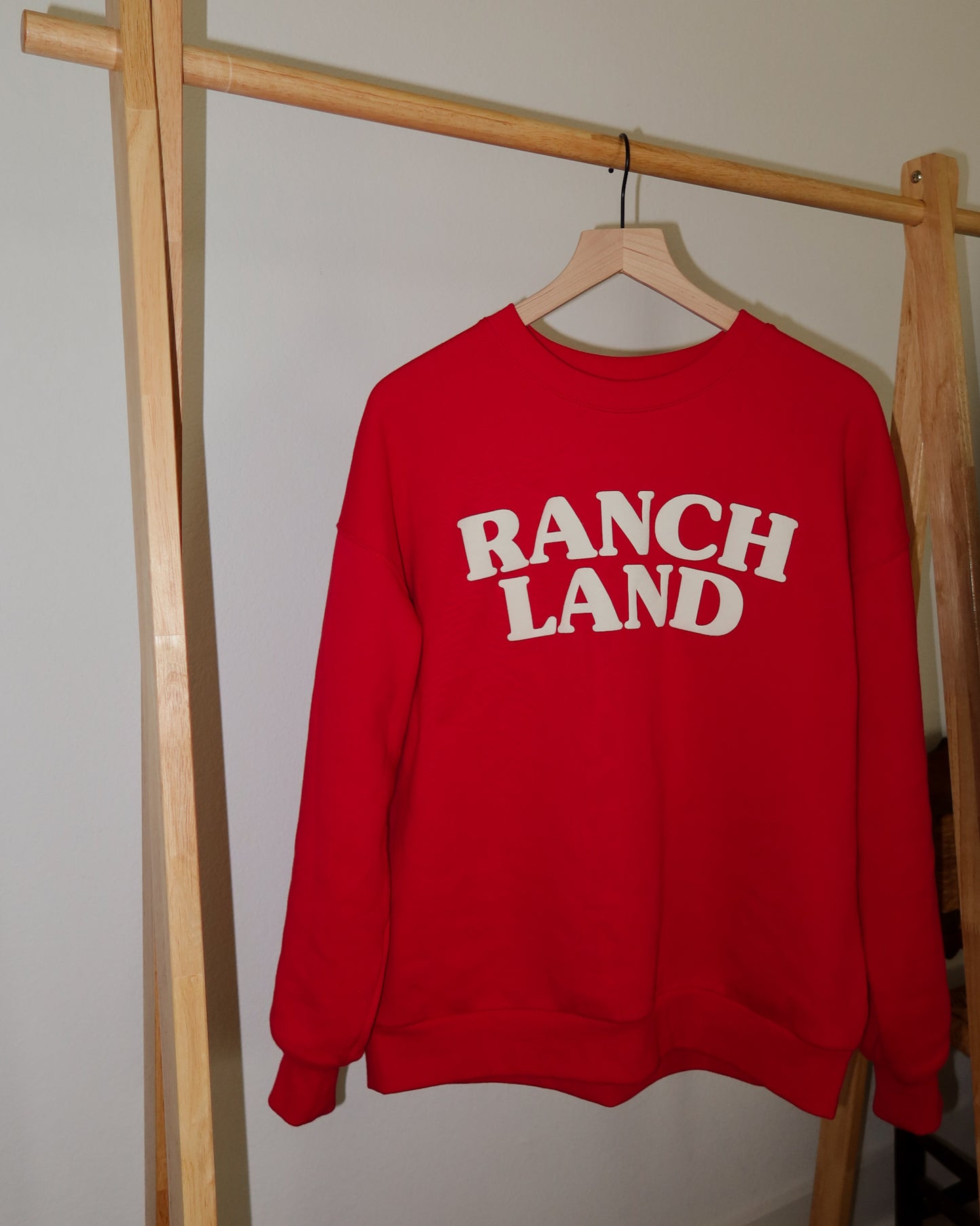 Red sweatshirt with 'RANCH LAND' text on a wooden hanger against a neutral wall.