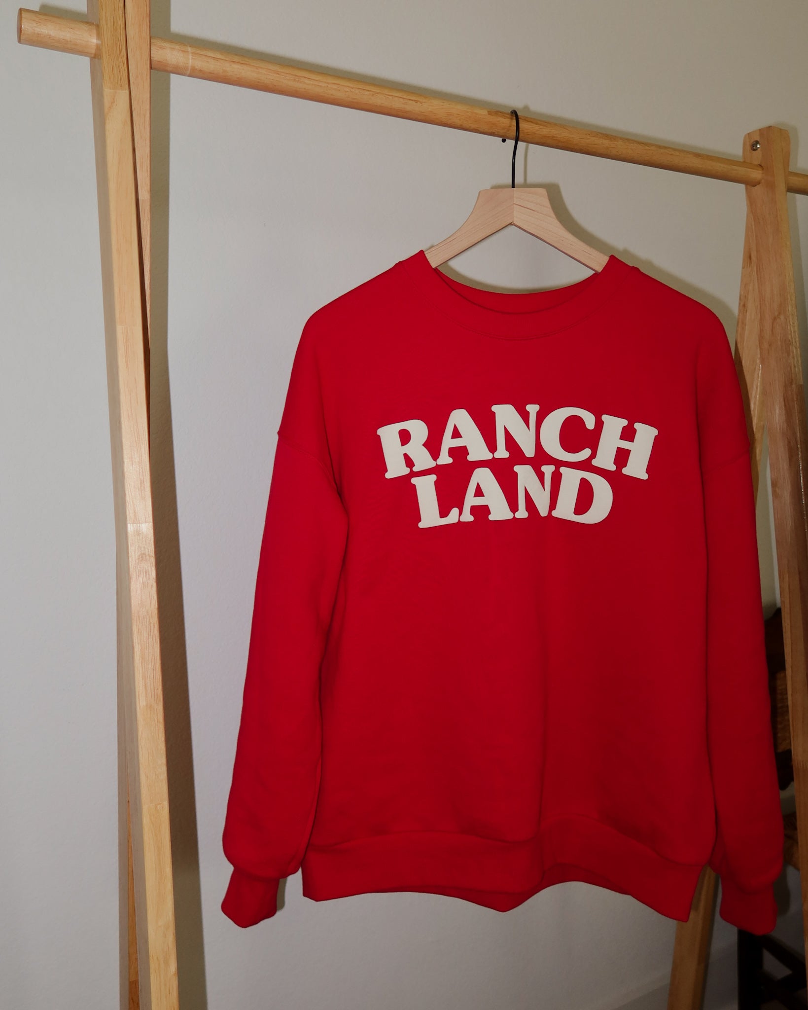 Red sweatshirt with 'RANCH LAND' text on a wooden hanger against a neutral wall.