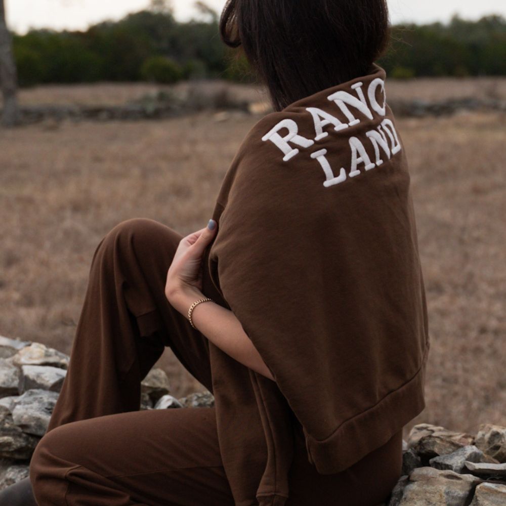 A model sits on a rustic stacked stone wall with her back to the camera, wearing a brown sweatshirt with 'RANCH LAND' embroidered in large white arched letters across the shoulders. She is wearing matching brown lounge pants.
