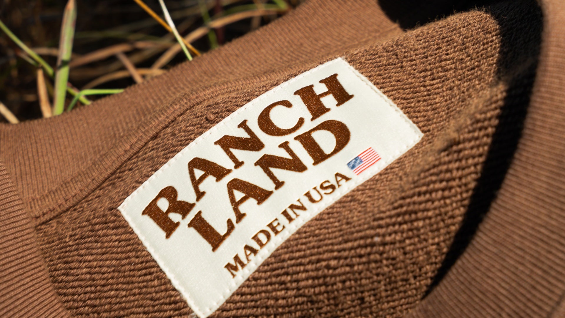 Close-up of a white woven clothing label on a brown ribbed neckline. The label features the brown 'RANCH LAND' logo, a small American flag, and the text 'MADE IN USA'.